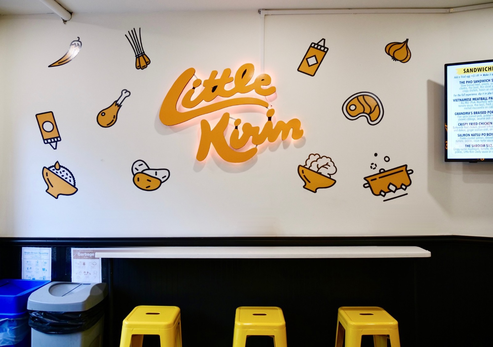 Little Kirin dining area: the glowing Little Kirin sign on a white wall with food doodles, a counter and yellow stools